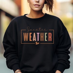 Puede incluir: Sudadera negra con la frase "Sweater Weather" en un marco rectangular. La palabra "WEATHER" est&aacute; en letras de colores. Un peque&ntilde;o coraz&oacute;n est&aacute; debajo del marco. Cuello redondo.