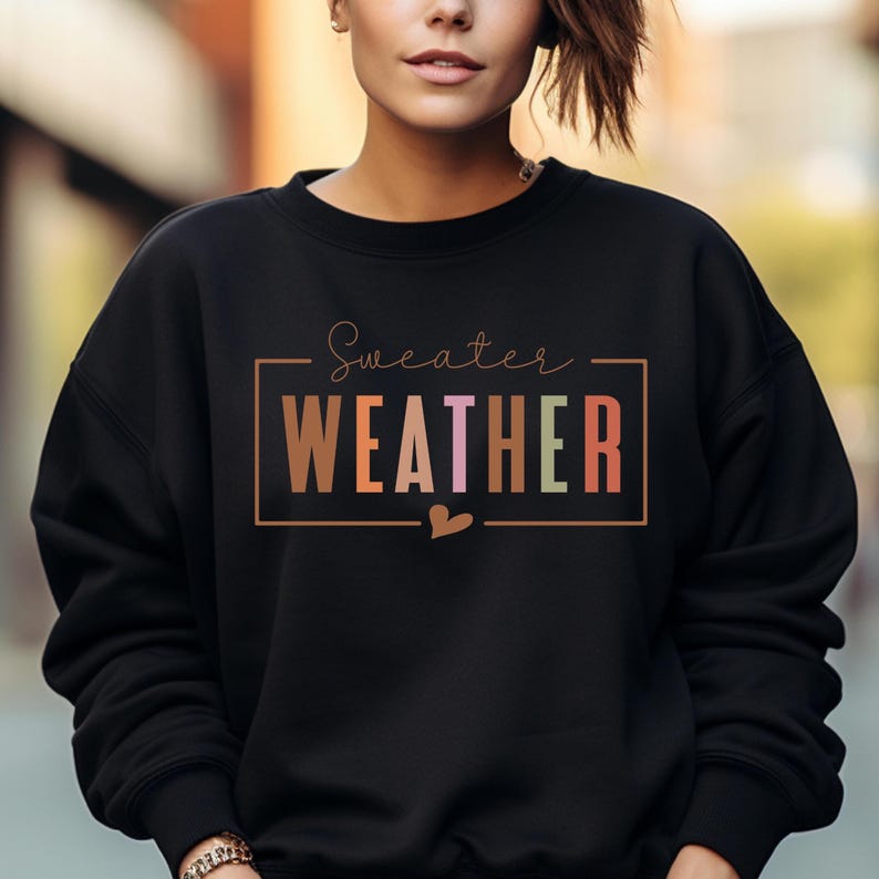 Puede incluir: Sudadera negra con la frase "Sweater Weather" en un marco rectangular. La palabra "WEATHER" est&aacute; en letras de colores. Un peque&ntilde;o coraz&oacute;n est&aacute; debajo del marco. Cuello redondo.