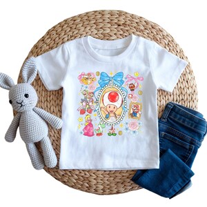 May include: White t-shirt featuring a colorful graphic of a cartoon character in an oval frame, surrounded by other characters and floral accents. A blue bow adorns the top of the frame. Dark blue jeans and a gray stuffed bunny are also in the image.