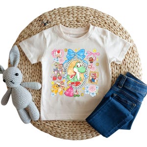 May include: A cream-colored t-shirt featuring a colorful graphic of Yoshi and other characters from the Super Mario video game series. The shirt is paired with blue denim jeans and a gray crocheted bunny toy.