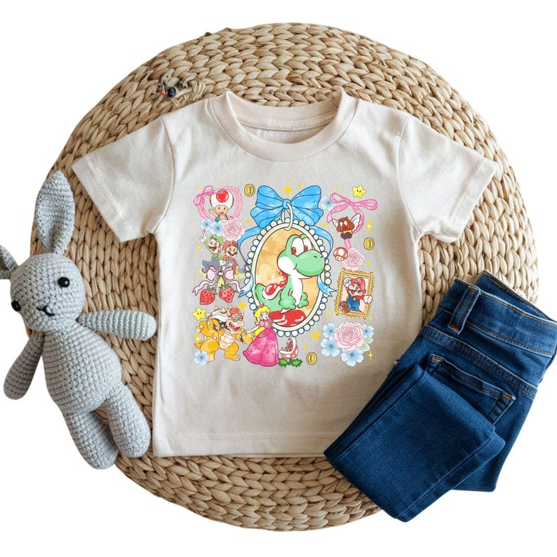 May include: A cream-colored t-shirt featuring a colorful graphic of Yoshi and other characters from the Super Mario video game series. The shirt is paired with blue denim jeans and a gray crocheted bunny toy.
