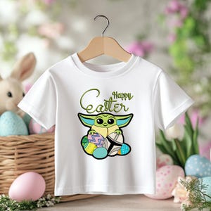 May include: White t-shirt featuring a cartoon character with large eyes and pointed ears, holding Easter eggs. The shirt has the words "Happy Easter" in green script. The shirt is hanging on a wooden hanger.