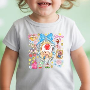 May include: A white toddler t-shirt featuring a colorful graphic of characters from the Super Mario video game series. The design includes Toad, Princess Peach, Mario, and other elements, surrounded by flowers and a blue bow.