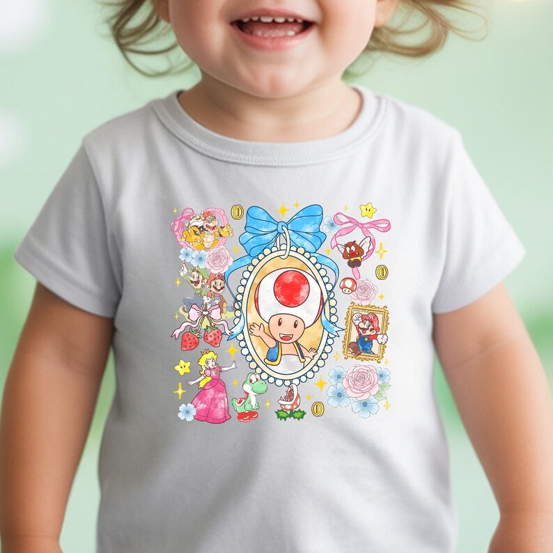 May include: A white toddler t-shirt featuring a colorful graphic of characters from the Super Mario video game series. The design includes Toad, Princess Peach, Mario, and other elements, surrounded by flowers and a blue bow.