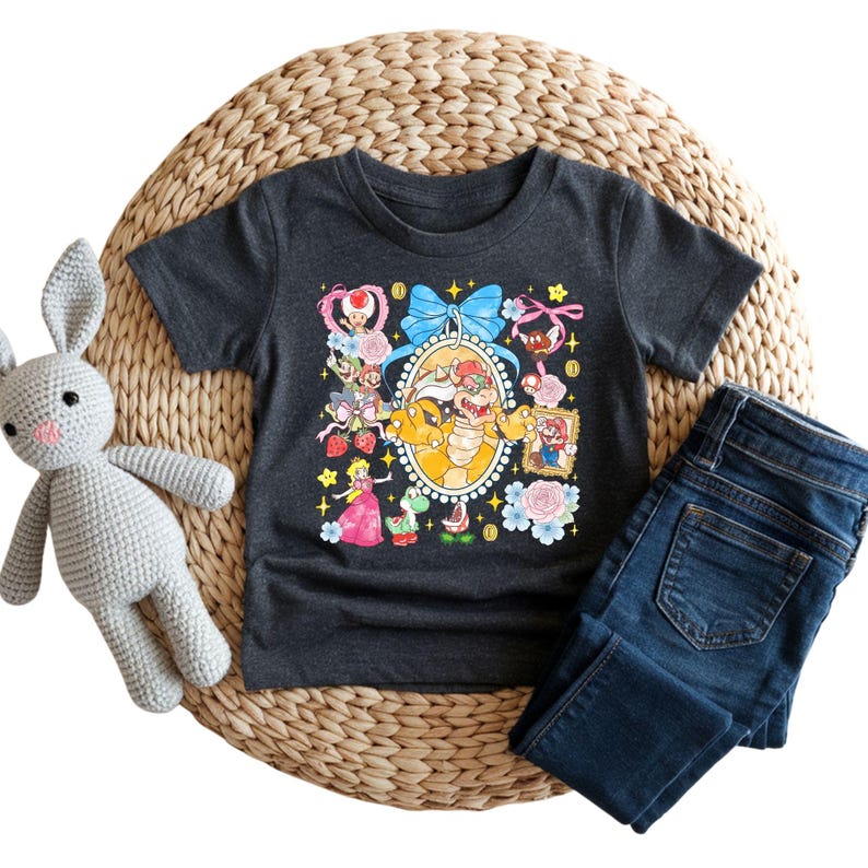 May include: Dark gray t-shirt featuring a colorful graphic of Bowser surrounded by other characters from the Super Mario Bros. universe. The shirt is paired with dark blue jeans and a gray stuffed bunny on a woven mat.