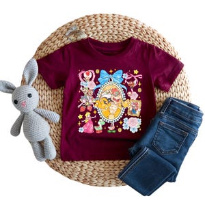 May include: Burgundy short-sleeve t-shirt featuring a colorful cartoon graphic of various characters. The graphic includes a large oval frame with a bow, flowers, and other decorative elements. Dark blue denim jeans are folded next to the shirt. A gray stuffed bunny is on the left.