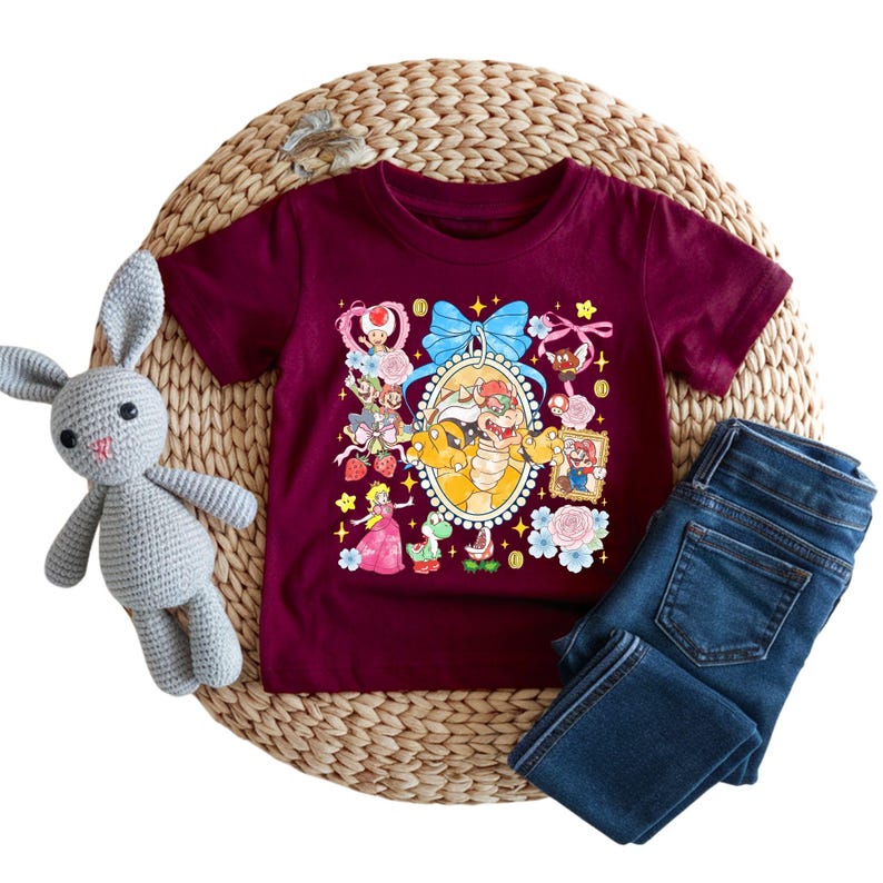 May include: Burgundy short-sleeve t-shirt featuring a colorful cartoon graphic of various characters. The graphic includes a large oval frame with a bow, flowers, and other decorative elements. Dark blue denim jeans are folded next to the shirt. A gray stuffed bunny is on the left.
