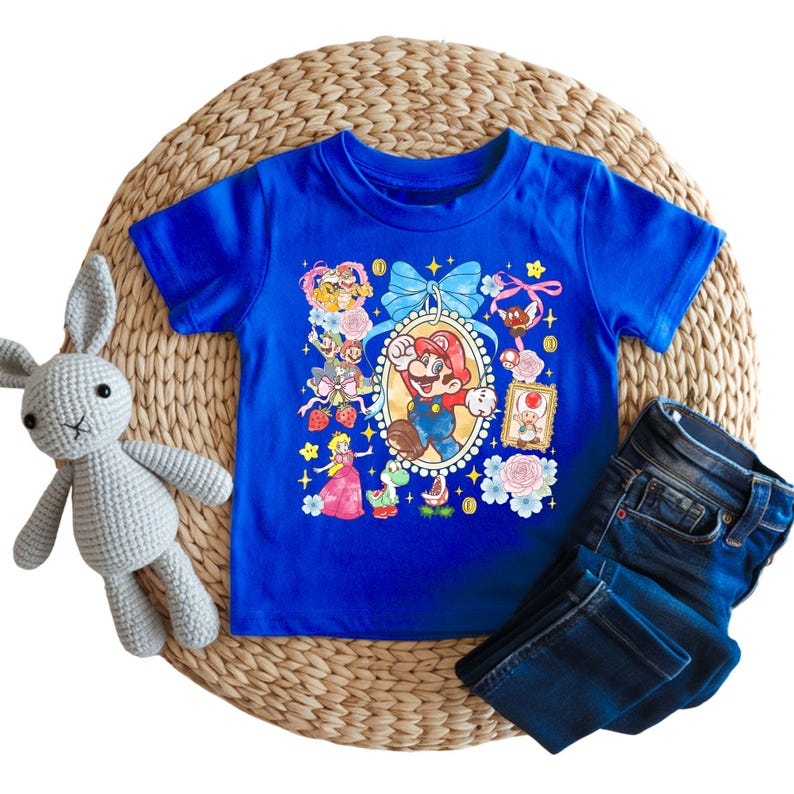 May include: A royal blue t-shirt featuring a colorful graphic of Mario and other characters. The shirt is paired with dark blue denim jeans and a gray crocheted bunny. The items are arranged on a woven mat.