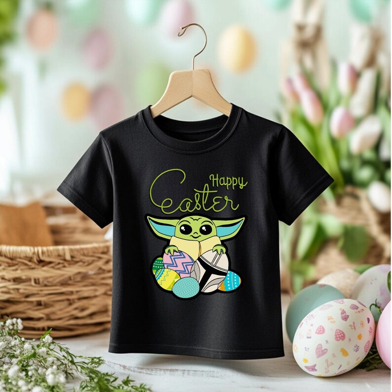 May include: Black t-shirt featuring a cartoon character with large ears and holding Easter eggs. The words "Happy Easter" are written in a script font above the character. The shirt is hanging on a wooden hanger.
