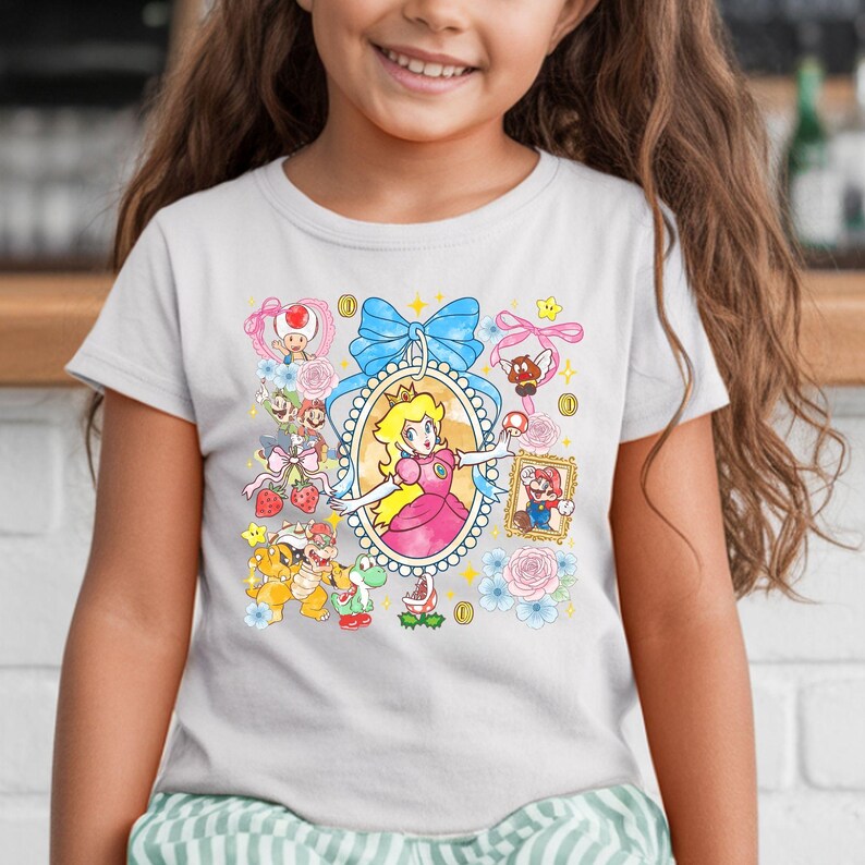 May include: White t-shirt featuring a colorful graphic of Princess Peach from the Super Mario video game series. The design includes various characters, coins, and floral elements, with a large blue bow at the top.