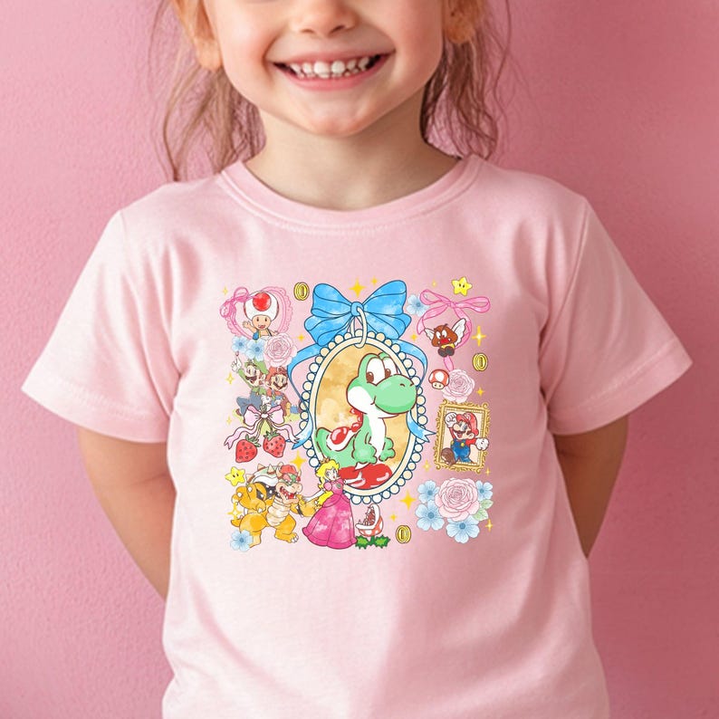 May include: A light pink t-shirt featuring a colorful graphic of Yoshi and other characters from the Super Mario video game series. The design includes a central oval frame with Yoshi, surrounded by flowers, bows, and other characters.