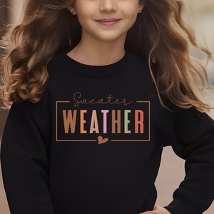 Puede incluir: Sudadera negra con la frase "Sweater Weather" en un marco rectangular. El texto utiliza un degradado de colores, incluyendo marr&oacute;n, naranja, rosa y verde. Un peque&ntilde;o coraz&oacute;n est&aacute; debajo del texto.
