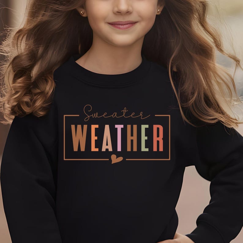 Puede incluir: Sudadera negra con la frase "Sweater Weather" en un marco rectangular. El texto utiliza un degradado de colores, incluyendo marr&oacute;n, naranja, rosa y verde. Un peque&ntilde;o coraz&oacute;n est&aacute; debajo del texto.