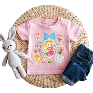 May include: A light pink t-shirt featuring a colorful graphic of Princess Peach from the Super Mario video game series. The design includes a portrait of Princess Peach, along with other characters and elements from the game. Dark blue denim jeans and a stuffed bunny are also in the image.