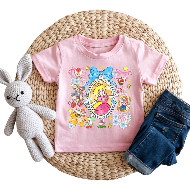 May include: A light pink t-shirt featuring a colorful graphic of Princess Peach from the Super Mario video game series. The design includes a portrait of Princess Peach, along with other characters and elements from the game. Dark blue denim jeans and a stuffed bunny are also in the image.