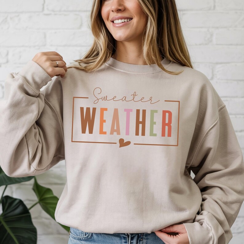 Puede incluir: Sudadera beige con la frase "Sweater Weather" en un dise&ntilde;o rectangular. La palabra "WEATHER" est&aacute; en letras de bloque marrones, naranjas, rosas y verdes. Un peque&ntilde;o coraz&oacute;n est&aacute; debajo del texto.