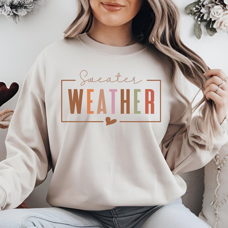 Puede incluir: Sudadera beige con la frase "Sweater Weather" en un marco rectangular. El texto es de varios colores: marr&oacute;n, naranja, rosa, verde y rojo. Un peque&ntilde;o coraz&oacute;n est&aacute; debajo del texto.