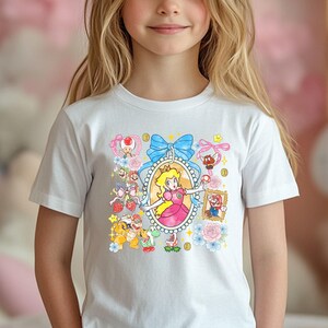 May include: White t-shirt featuring a colorful graphic of Princess Peach in an oval frame, surrounded by other Super Mario characters, blue bows, and flowers. The design includes Mario, Toad, Yoshi, and coins.
