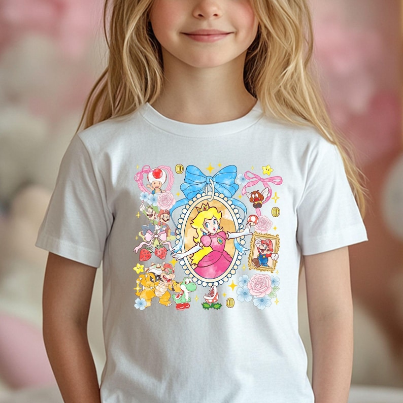May include: White t-shirt featuring a colorful graphic of Princess Peach in an oval frame, surrounded by other Super Mario characters, blue bows, and flowers. The design includes Mario, Toad, Yoshi, and coins.