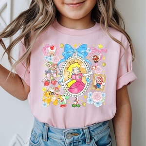 May include: Pink t-shirt featuring a colorful graphic of Princess Peach surrounded by other characters and elements from the Super Mario video game series. The design includes a blue bow, flowers, and gold coins.