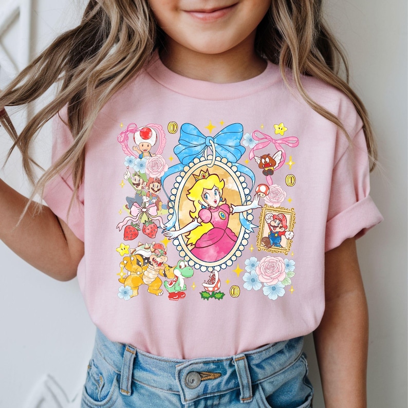 May include: Pink t-shirt featuring a colorful graphic of Princess Peach surrounded by other characters and elements from the Super Mario video game series. The design includes a blue bow, flowers, and gold coins.