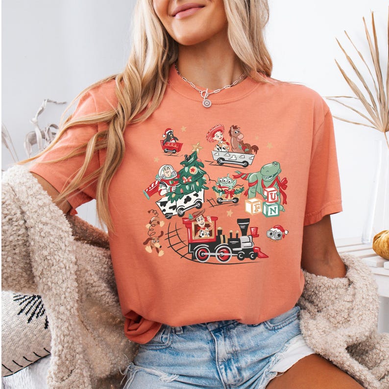 May include: Coral-colored t-shirt featuring a festive graphic with cartoon characters, including a train, Christmas tree, and various toys. The design incorporates elements of a holiday theme, with a playful and colorful aesthetic.