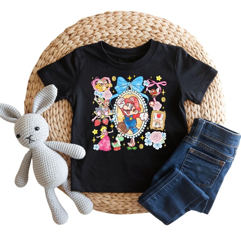 May include: Black t-shirt featuring a colorful graphic of Mario and other characters. The design includes a blue bow, flowers, and a framed image of Mario. Paired with blue jeans and a gray stuffed bunny.