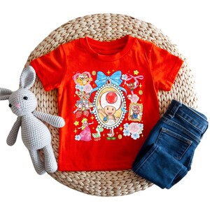 May include: A red t-shirt featuring a colorful graphic of characters from the Super Mario video game series. The design includes Toad, Princess Peach, and Mario. The shirt is paired with blue denim jeans and a gray stuffed bunny.