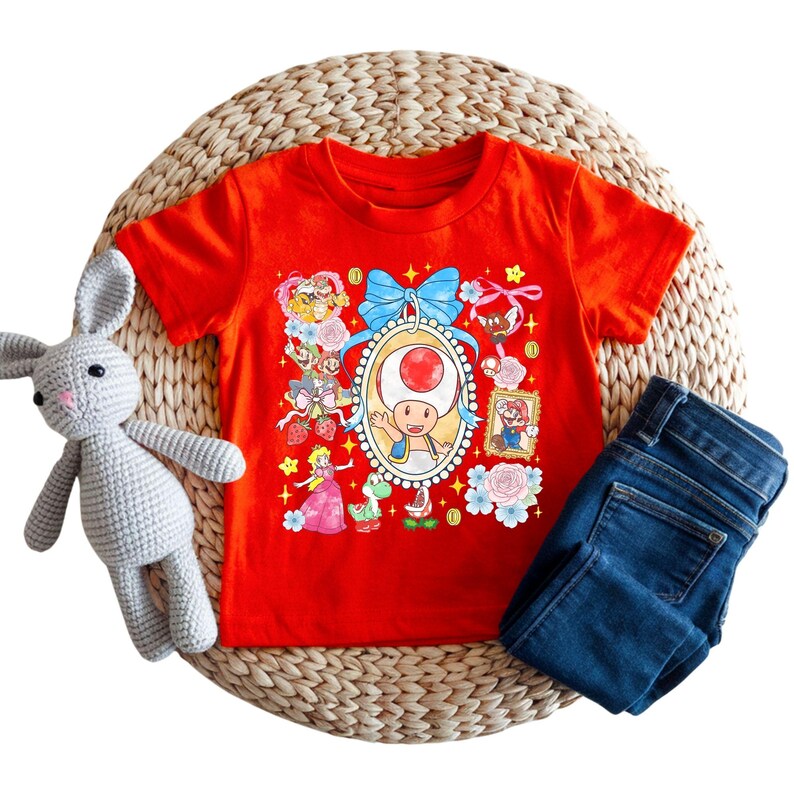 May include: A red t-shirt featuring a colorful graphic of characters from the Super Mario video game series. The design includes Toad, Princess Peach, and Mario. The shirt is paired with blue denim jeans and a gray stuffed bunny.
