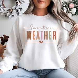 Puede incluir: Sudadera blanca con la frase "Sweater Weather" en un dise&ntilde;o rectangular. La palabra "WEATHER" est&aacute; en marr&oacute;n, rosa y verde. Un peque&ntilde;o coraz&oacute;n marr&oacute;n est&aacute; debajo. La sudadera es de estilo casual.