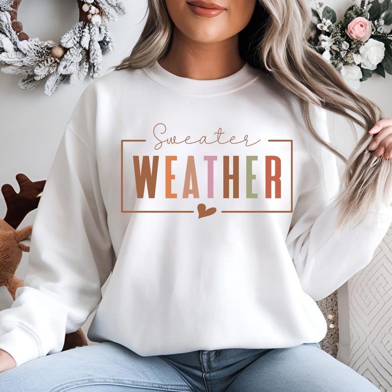 Puede incluir: Sudadera blanca con la frase "Sweater Weather" en un dise&ntilde;o rectangular. La palabra "WEATHER" est&aacute; en marr&oacute;n, rosa y verde. Un peque&ntilde;o coraz&oacute;n marr&oacute;n est&aacute; debajo. La sudadera es de estilo casual.