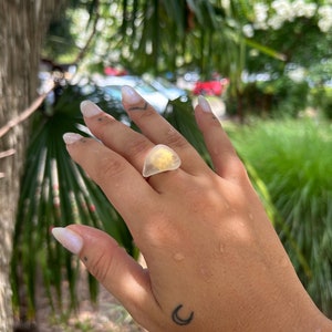 May include: A clear resin ring with a yellow flower inside. The ring is on a person's finger. The person has a crescent moon tattoo on their hand.