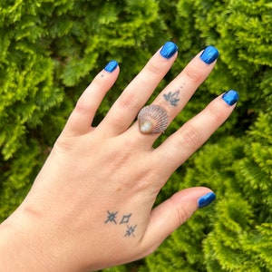 Peut inclure: Une main avec du vernis à ongles bleu porte une bague avec une coquille de mer. La main a un petit tatouage de trois étoiles sur la paume.