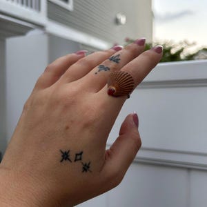 Puede incluir: Una mano con un pequeño tatuaje de tres estrellas en la palma, llevando un anillo hecho de una concha marrón.