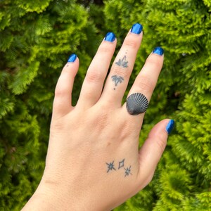 Könnte beinhalten: Eine Hand mit blauem Nagellack trägt einen silbernen Ring mit einem schwarzen Muschel-Design. Die Hand hat mehrere kleine Tattoos, darunter Sterne und eine Blume.