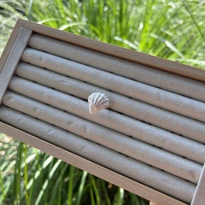 Puede incluir: Una caja de madera para exhibir anillos con un interior de terciopelo blanco y múltiples ranuras para anillos. Una pequeña concha blanca se coloca en una de las ranuras.