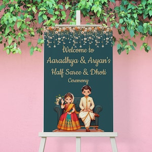 May include: Welcome to Aaradhya & Aryan's Half Saree & Dhoti Ceremony sign featuring a cartoon illustration of a girl and boy in traditional Indian clothing. The sign is on a wooden easel against a pink wall.