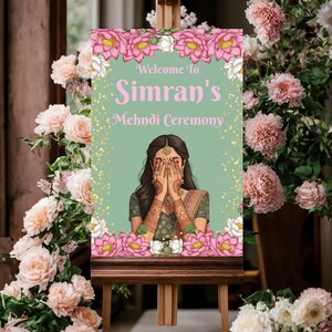 May include: A wooden easel displays a sign with the text "Welcome to Simran's Mehndi Ceremony." The sign features a woman with hennaed hands and lotus flower accents. Pink and white flowers frame the sign.