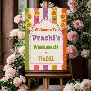 Puede incluir: Un letrero decorativo sobre un caballete de madera con el texto "Welcome to Prachi's Mehendi & Haldi". El letrero tiene un diseño colorido con patrones florales y a rayas. Flores rosas y blancas rodean el letrero.