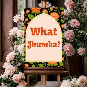 Op de afbeelding: Een houten ezel toont een bord met de tekst "What Jhumka?" in oranje. Het bord heeft een zwarte achtergrond met oranje bloempatronen en een crèmekleurig middenpaneel. De ezel is omgeven door roze en witte bloemen.