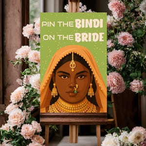 May include: A wooden easel displays a sign with the text "Pin the Bindi on the Bride." The sign features an illustration of a woman in traditional Indian attire, with a green background. The scene is framed by pink flowers.