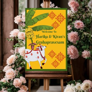 May include: A welcome sign for Harika & Kiran's Gruhapravesam, featuring a yellow background with green accents and a white cow illustration. The sign is displayed on a wooden easel, surrounded by pink flowers.