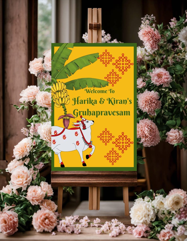 May include: A welcome sign for Harika & Kiran's Gruhapravesam, featuring a yellow background with green accents and a white cow illustration. The sign is displayed on a wooden easel, surrounded by pink flowers.