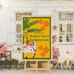 May include: A wooden easel displays a sign with a yellow background and green border. The sign features text that reads "Welcome to Harika & Kiran's Gruhapravesam." A white cow illustration with red accents is on the sign.