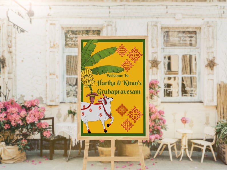 May include: A wooden easel displays a sign with a yellow background and green border. The sign features text that reads "Welcome to Harika & Kiran's Gruhapravesam." A white cow illustration with red accents is on the sign.