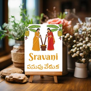 May include: A small wooden easel displays a white sign with a colorful illustration of two women in traditional Indian attire. The sign features the word "Sravani" in gold lettering, along with text in another language. The background is blurred with floral arrangements.