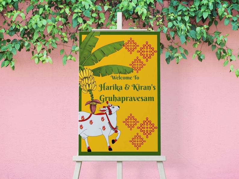 May include: A welcome sign for Harika & Kiran's Gruhapravesam, featuring a yellow background with green accents. The sign includes a drawing of a white cow with red details, banana leaves, and text. The sign is displayed on a white easel.
