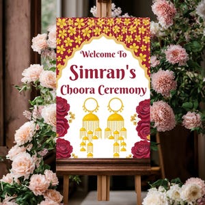 May include: A wooden easel displays a sign with the text "Welcome To Simran's Choora Ceremony." The sign features a maroon background with gold floral patterns and illustrations of gold earrings. Pink and red roses and flowers surround the sign.