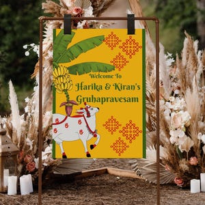 May include: A decorative sign with the text "Welcome to Harika & Kiran's Gruhapravesam" in green and gold. The sign features a white cow illustration with red accents, bananas, and green leaves. The sign is displayed on a brown metal stand.