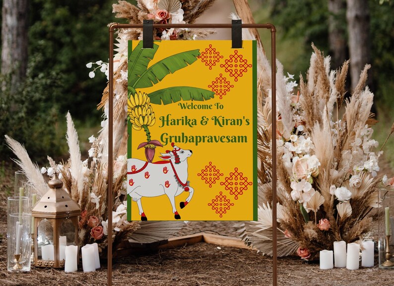 May include: A decorative sign with the text "Welcome to Harika & Kiran's Gruhapravesam" in green and gold. The sign features a white cow illustration with red accents, bananas, and green leaves. The sign is displayed on a brown metal stand.